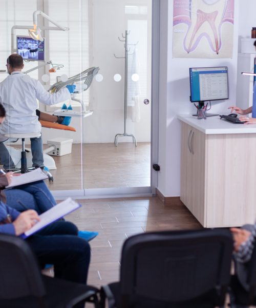 Crowded stomatology waiting area with people filling form for dental consultation. Stomatoloy specialist denstiry treating senior woman cavity. Receptionist working on computer.